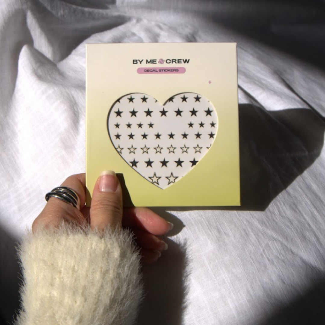 Hand holding a card with a heart and star design, branded 'By Me & Crew'.